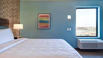 Una habitación de hotel con cama, mesita de noche con lámpara, aire acondicionado instalado en la pared, ventana con vista a la ciudad y un cuadro abstracto enmarcado.