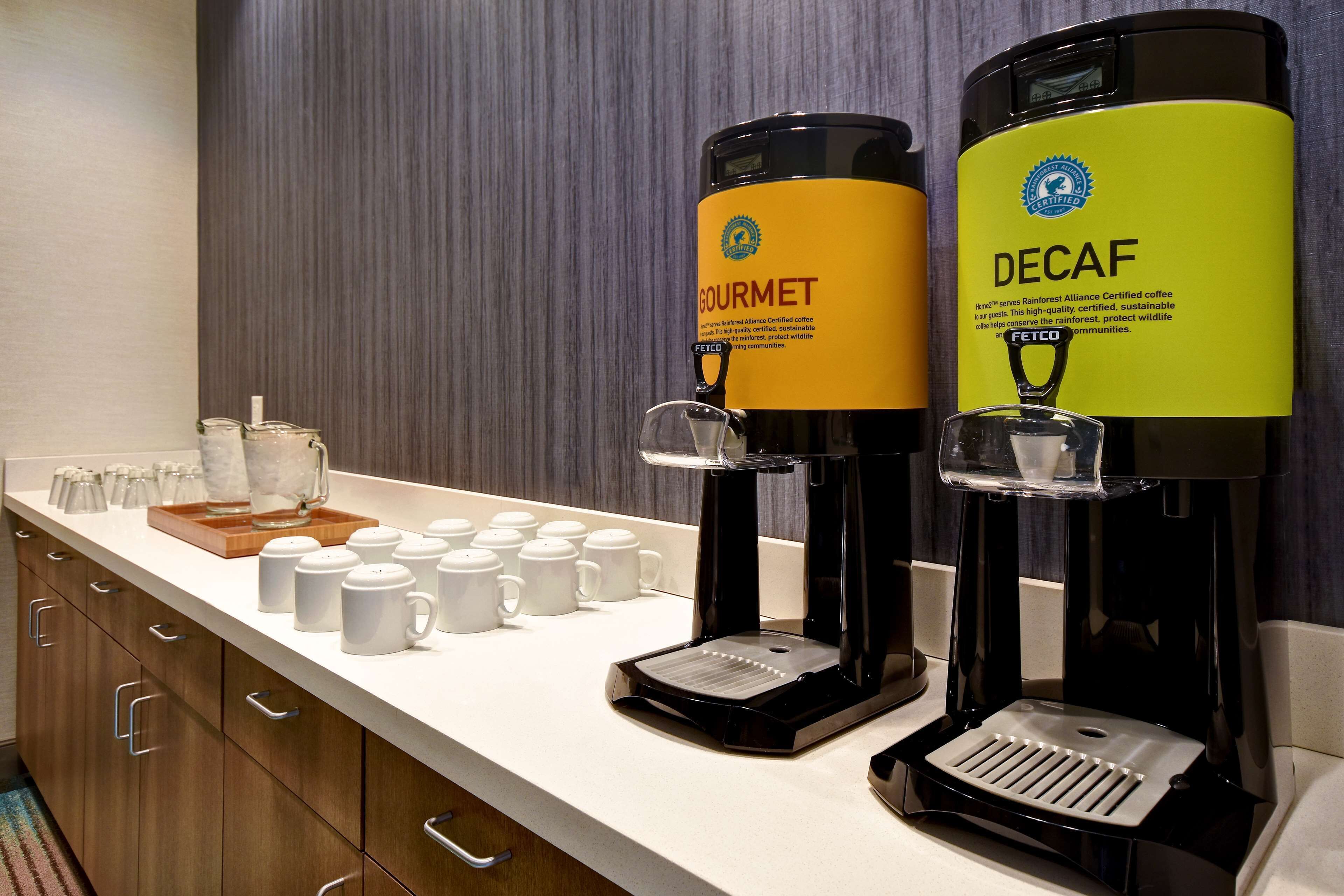 Una estación de café con dos dispensadores de café, uno etiquetado 'GOURMET' y otro 'DECAF', una bandeja con tazas de café y un molinillo.