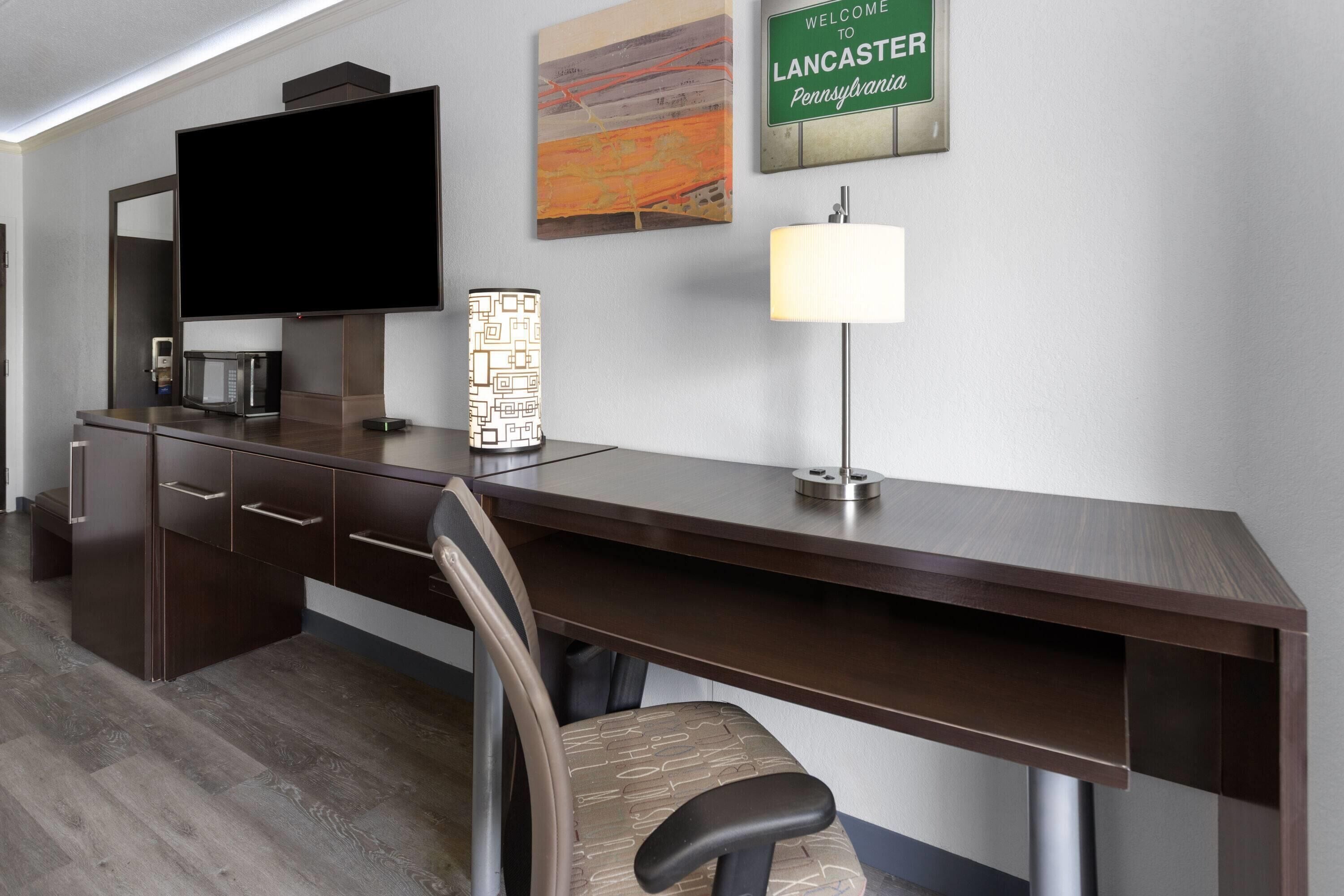Une chambre d’hôtel moderne équipée d’un grand téléviseur à écran plat, d’un bureau avec une lampe, d’un four à micro-ondes et d’un élément décoratif mural.