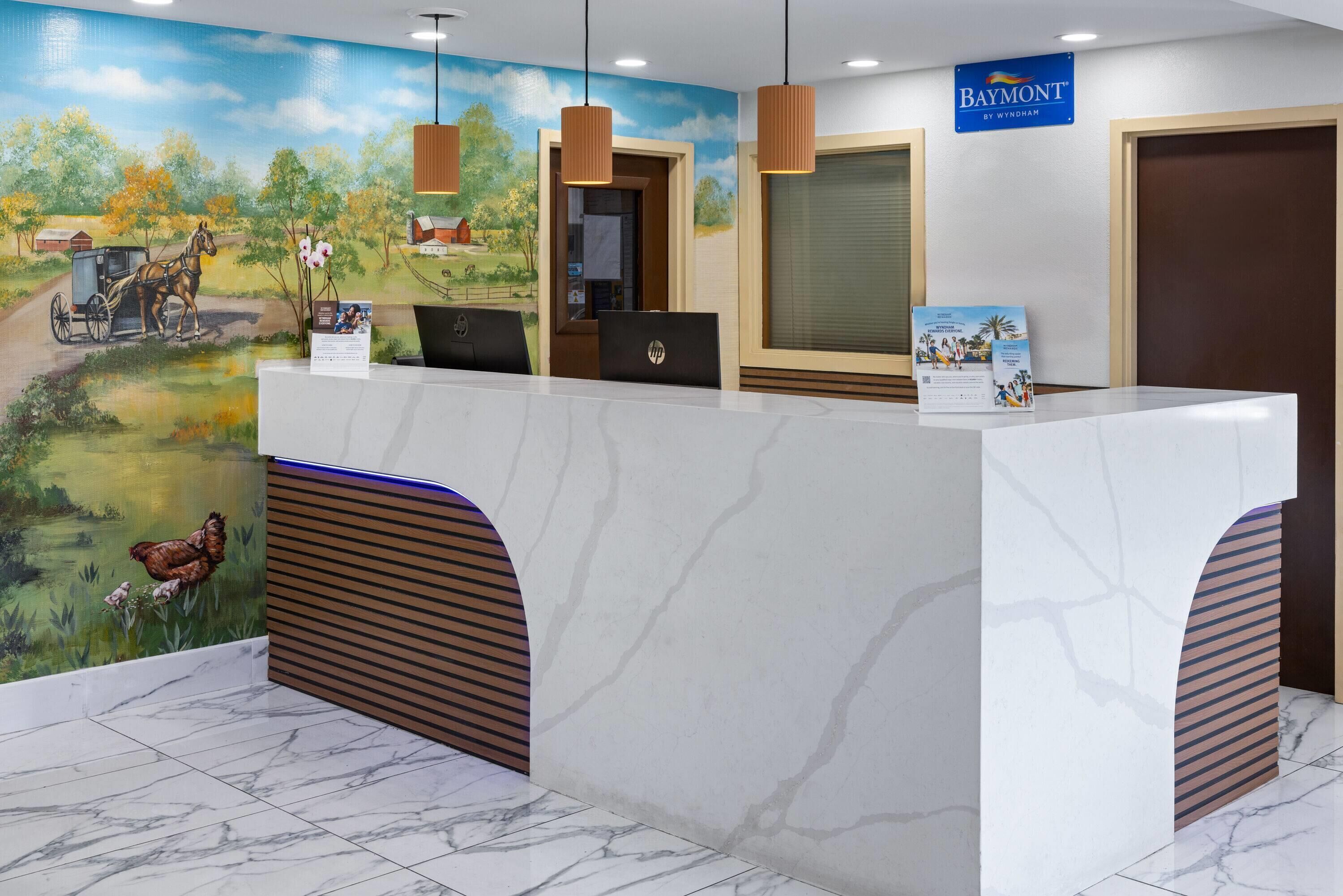A reception desk with a mural of a rural landscape, a sign for Baymont by Wyndham, and a laptop on the counter.