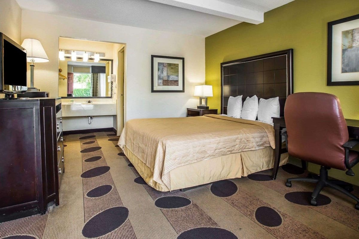 A hotel room with a bed, a desk, a chair, a mirror, a nightstand, and a picture on the wall.