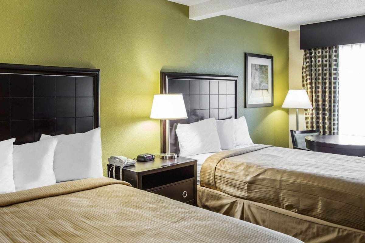 A hotel room with two beds, a nightstand with a phone and a lamp, and a window with curtains.