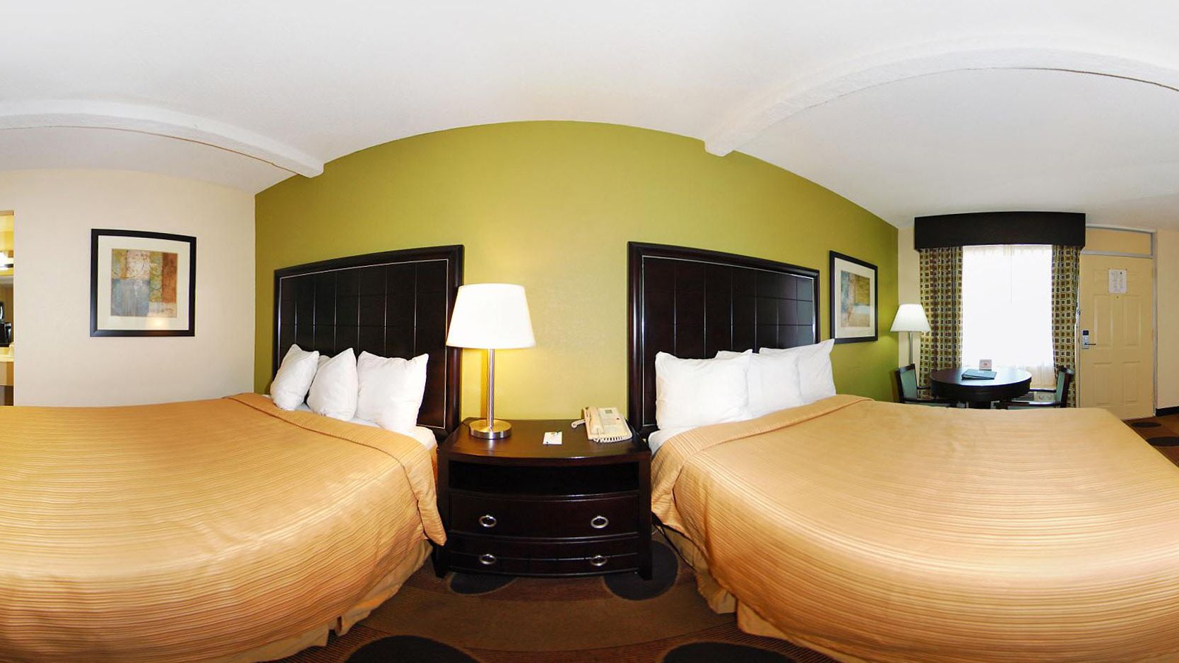 A hotel room with two beds, a nightstand with a lamp, and a small table with a chair.
