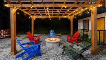 Un espacio al aire libre con una fogata, sillas de madera y una pérgola con luces colgantes.