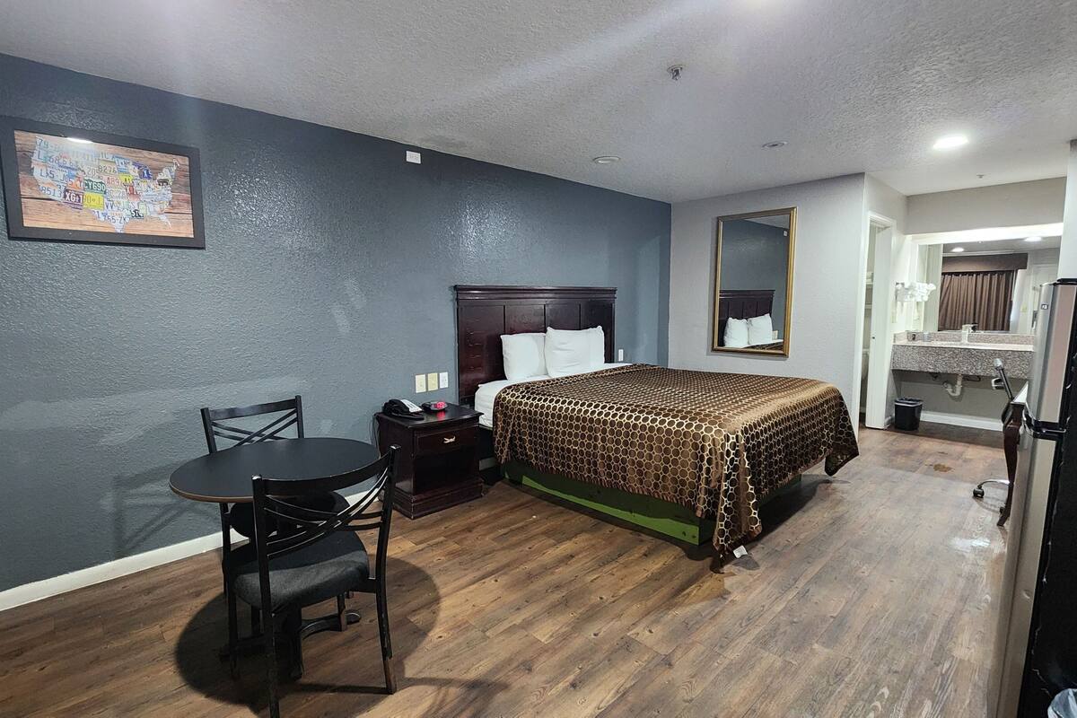 A hotel room with a bed, a desk with a chair, a small table, a mirror, and a refrigerator.
