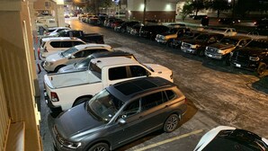 Un estacionamiento con varios coches aparcados en filas, un edificio de techo rojo y una calle con farolas.