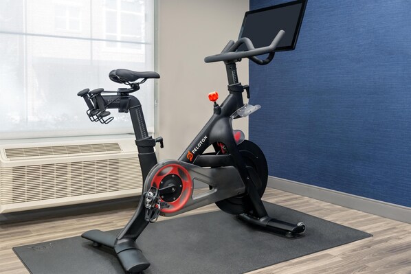 A stationary bike in a room with a large window, a wall-mounted screen, and a mat on the floor.