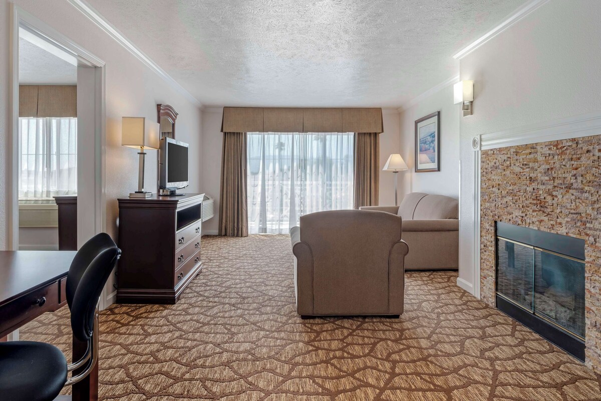 A hotel room with a patterned carpet, a sofa, a TV on a stand, a desk with a chair, a dresser, and a fireplace.
