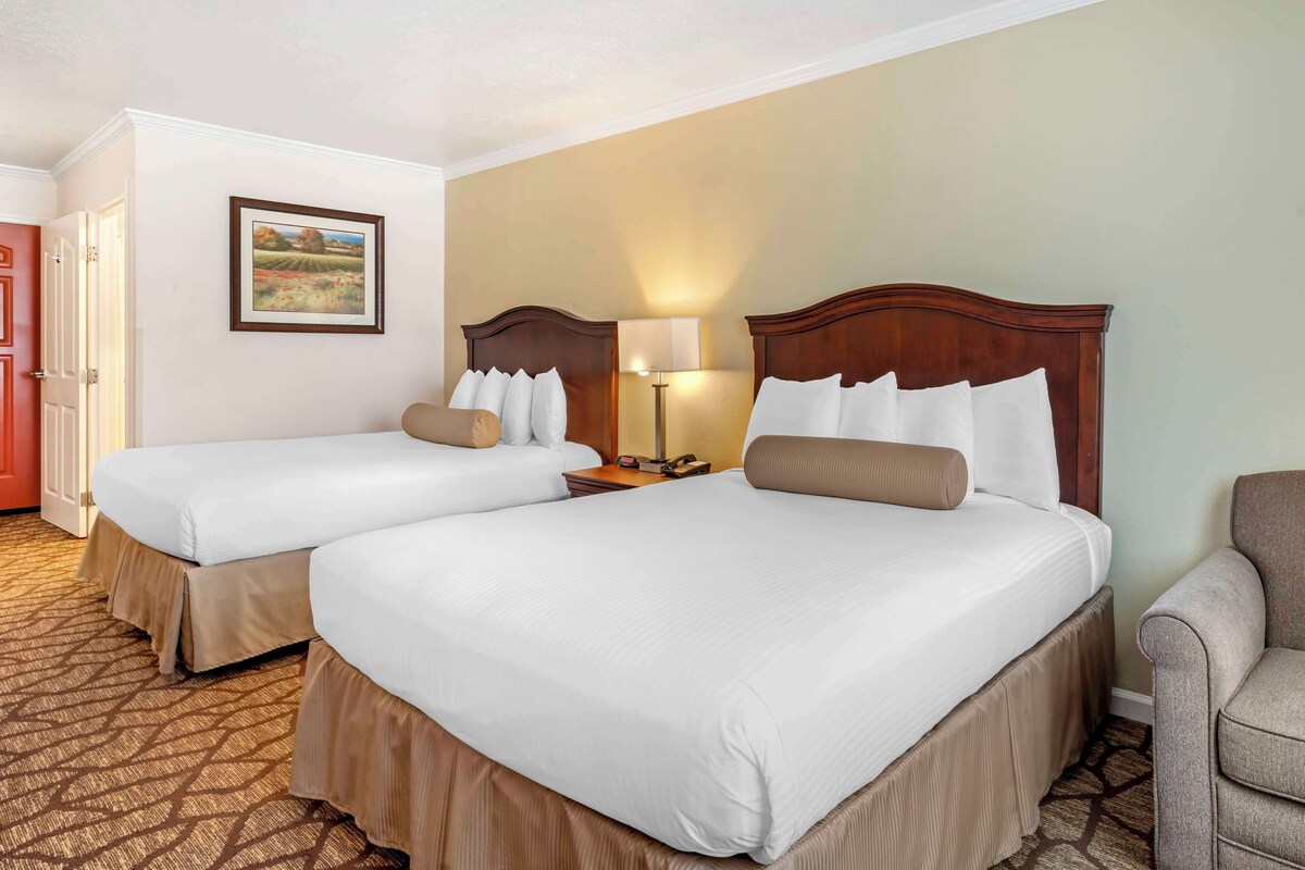 A hotel room with two beds, a nightstand, a lamp, and a chair.