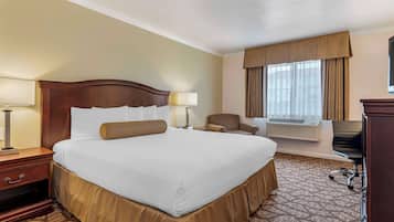 A hotel room with a large bed, a desk, a chair, a desk lamp, a window with curtains, and an air conditioning unit.