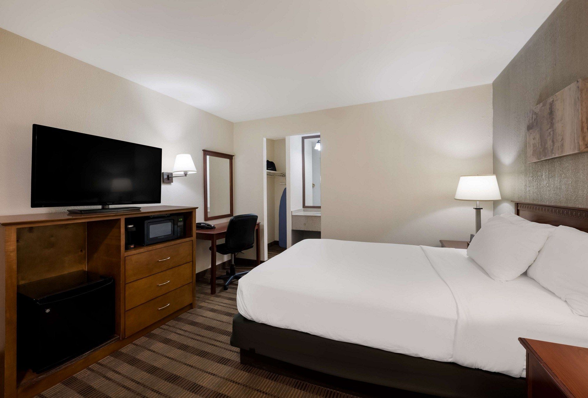A hotel room with a bed, a desk with a television, a chair, a desk lamp, and a bedside table.