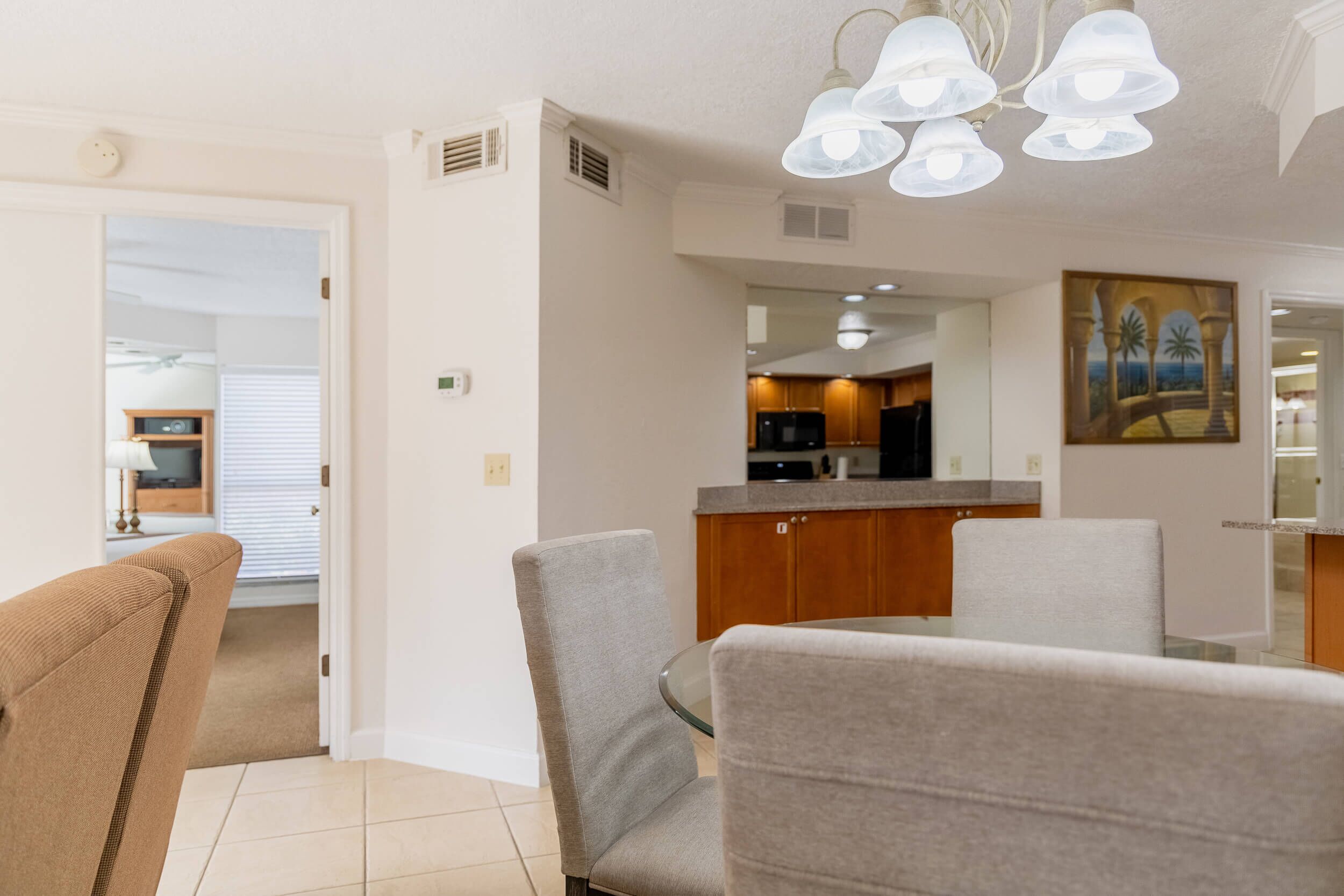 A dining area with a glass table and chairs, a kitchen in the background, and a ceiling light fixture.