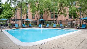 Una piscina rectangular con un borde empedrado, rodeada de sillas de descanso y sombrillas, y un edificio de ladrillo al fondo.