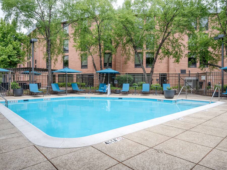 Una piscina rectangular con un borde empedrado, rodeada de sillas de descanso y sombrillas, y un edificio de ladrillo al fondo.
