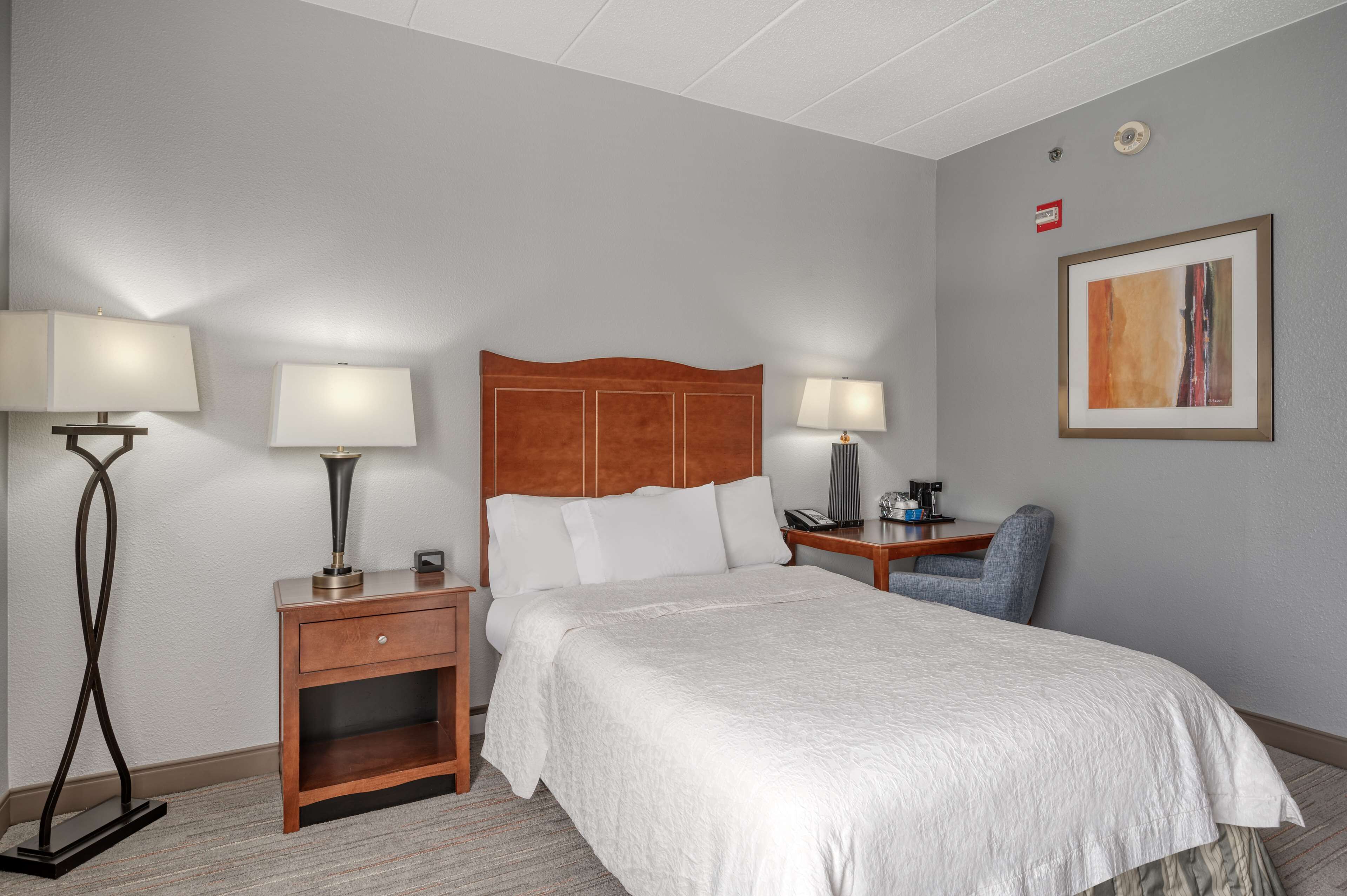 A hotel room with a bed, two bedside tables, a chair, a lamp, and a framed picture on the wall.