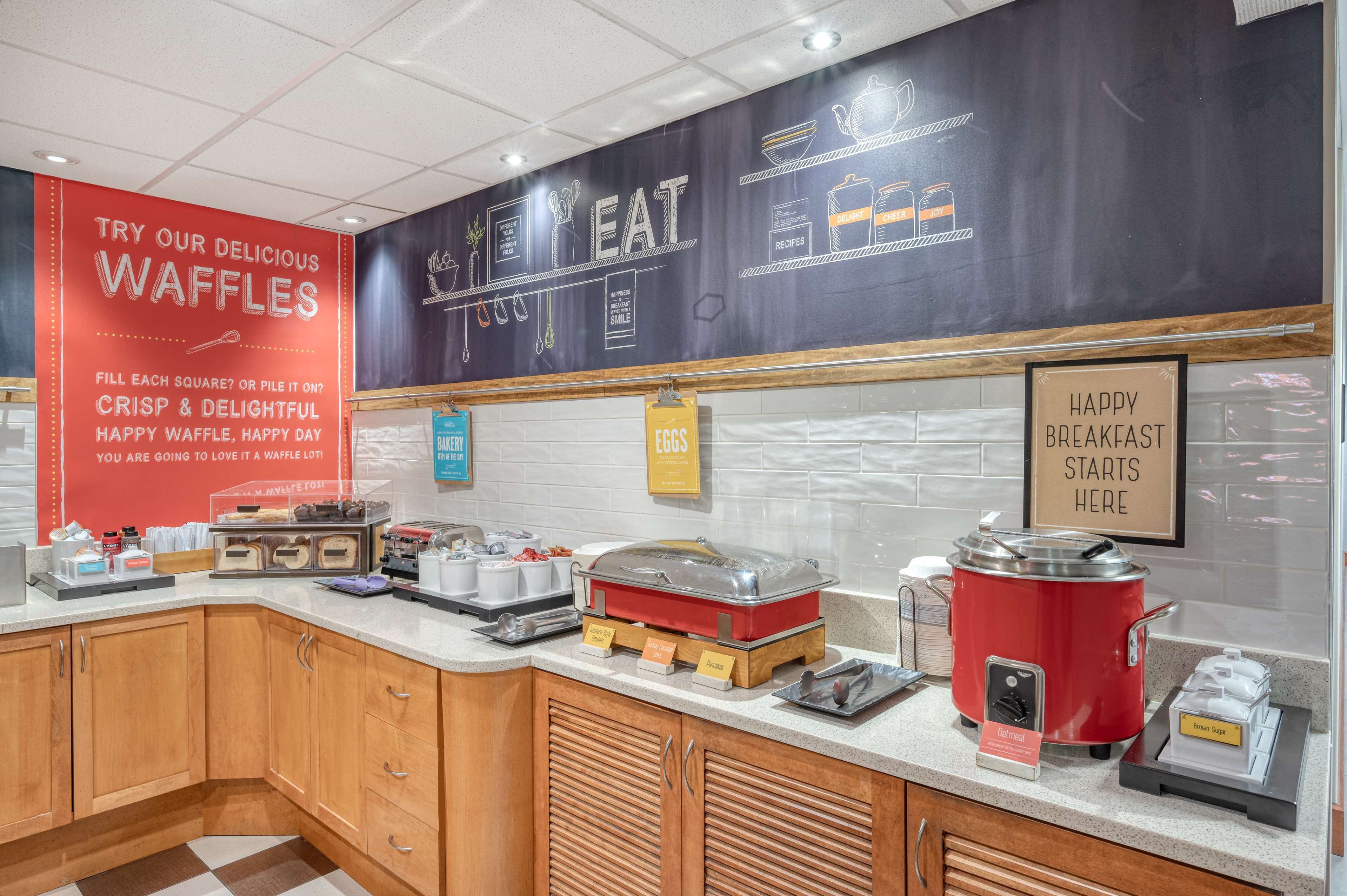 Zona de desayuno tipo buffet con un letrero rojo que anuncia gofres, un menú en pizarra y diversas opciones para el desayuno, como huevos y jarabe.