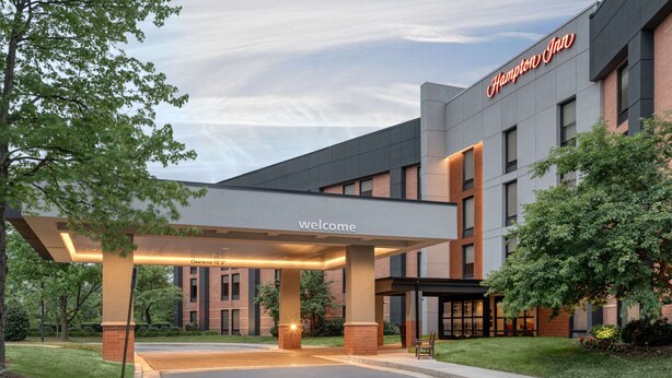 Un Hampton Inn con un letrero que dice 'Welcome' bajo un acceso cubierto, rodeado de follaje.