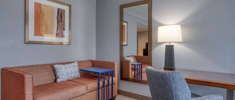 Habitación de hotel con sofá, una mesita, una silla, escritorio, lámpara y un cuadro en la pared.