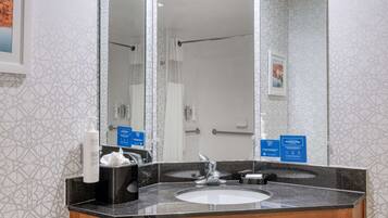 Un baño con un espejo grande, un lavamanos y una ducha. Hay letreros en la pared y una botella de jabón de manos sobre el mostrador.