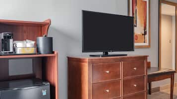 A hotel room with a flat-screen TV, a coffee maker, a mini-fridge, a wooden dresser, and a framed artwork on the wall.