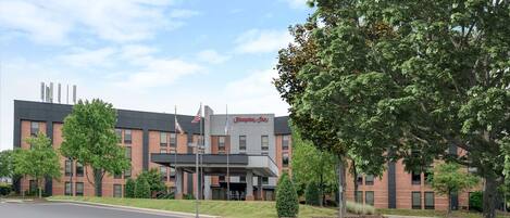 Un hotel Hampton Inn con fachada de ladrillo, una bandera y un césped cuidado.