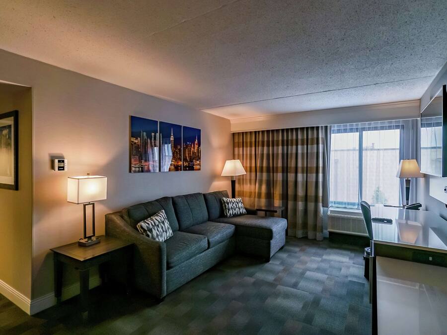 A hotel room with a sofa, two side tables with lamps, a flat-screen TV, a window with curtains, and a cityscape painting on the wall.