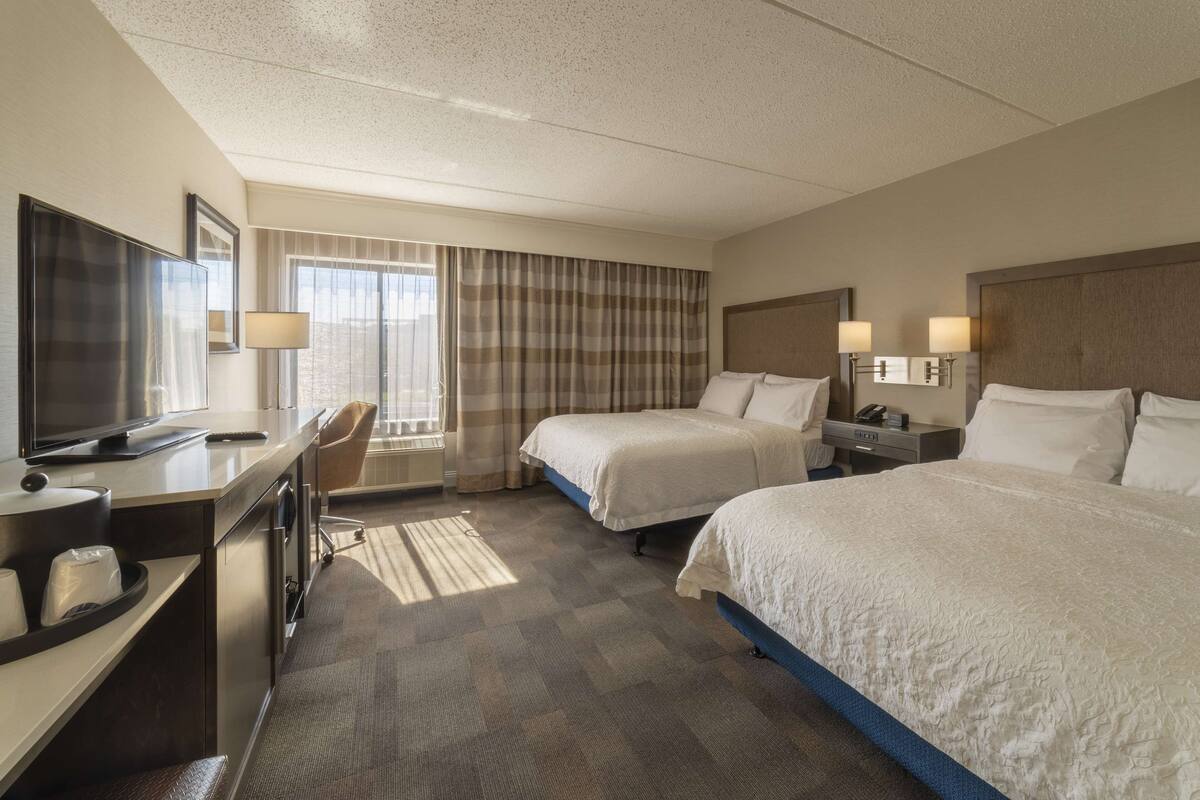 Une chambre d’hôtel avec deux lits, un bureau avec une télévision, une chaise et une fenêtre avec des rideaux.