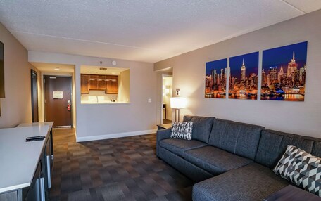 A modern hotel room with a sofa, a dining table, a kitchen area, and cityscape artwork on the wall.
