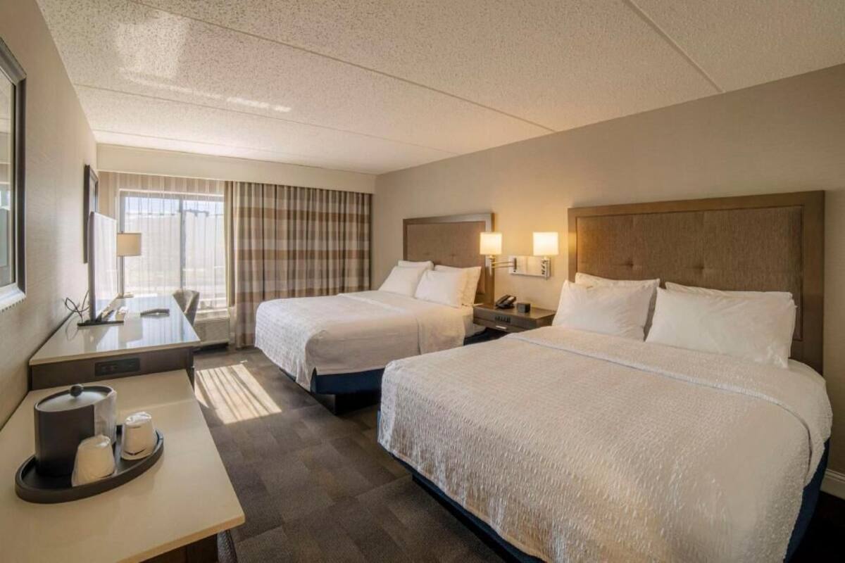 Une chambre d’hôtel avec deux lits, un bureau avec une lampe, une fenêtre avec des rideaux et un plateau avec des tasses et un récipient.