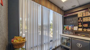 Un área de café con una pizarra que muestra '¡Disfruta tu día!!' y varios aperitivos y bebidas en estantes.