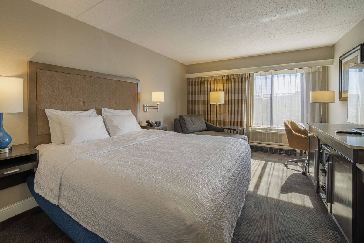 Une chambre d’hôtel avec un grand lit, un bureau avec une chaise, une lampe de bureau, une chaise, une fenêtre avec des rideaux et un miroir.