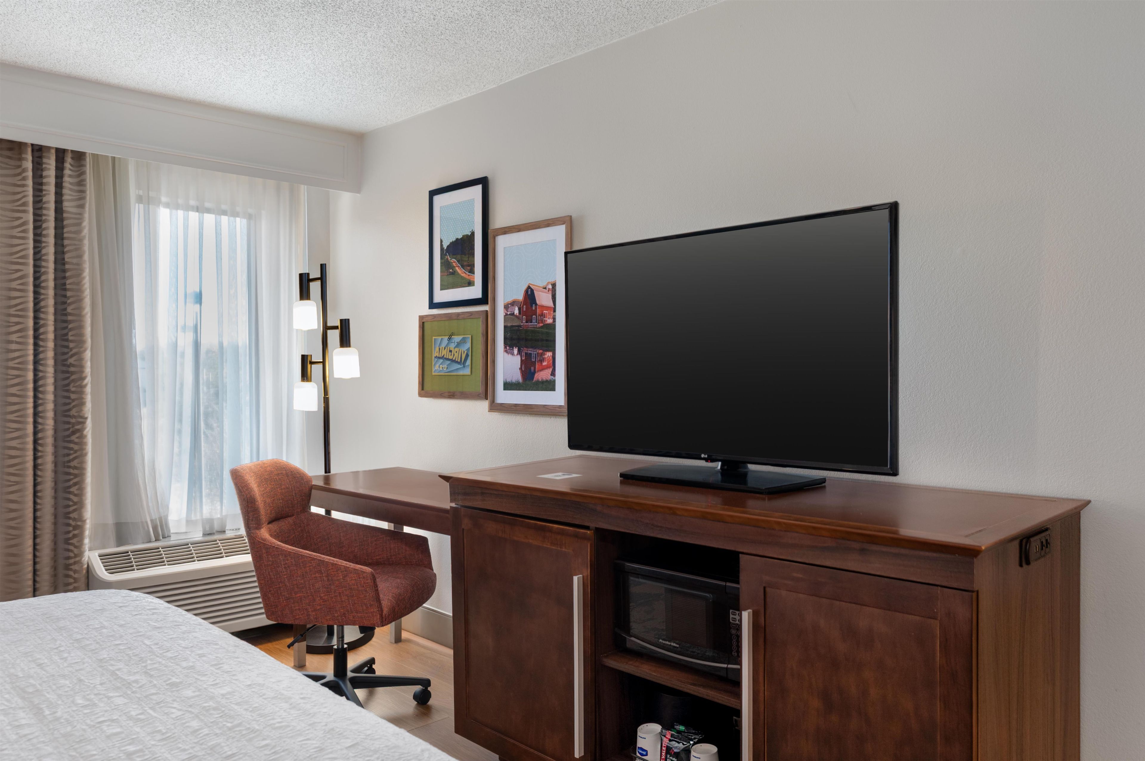 A hotel room with a bed, a desk with a flat-screen TV, a chair, a microwave, and a window with curtains.