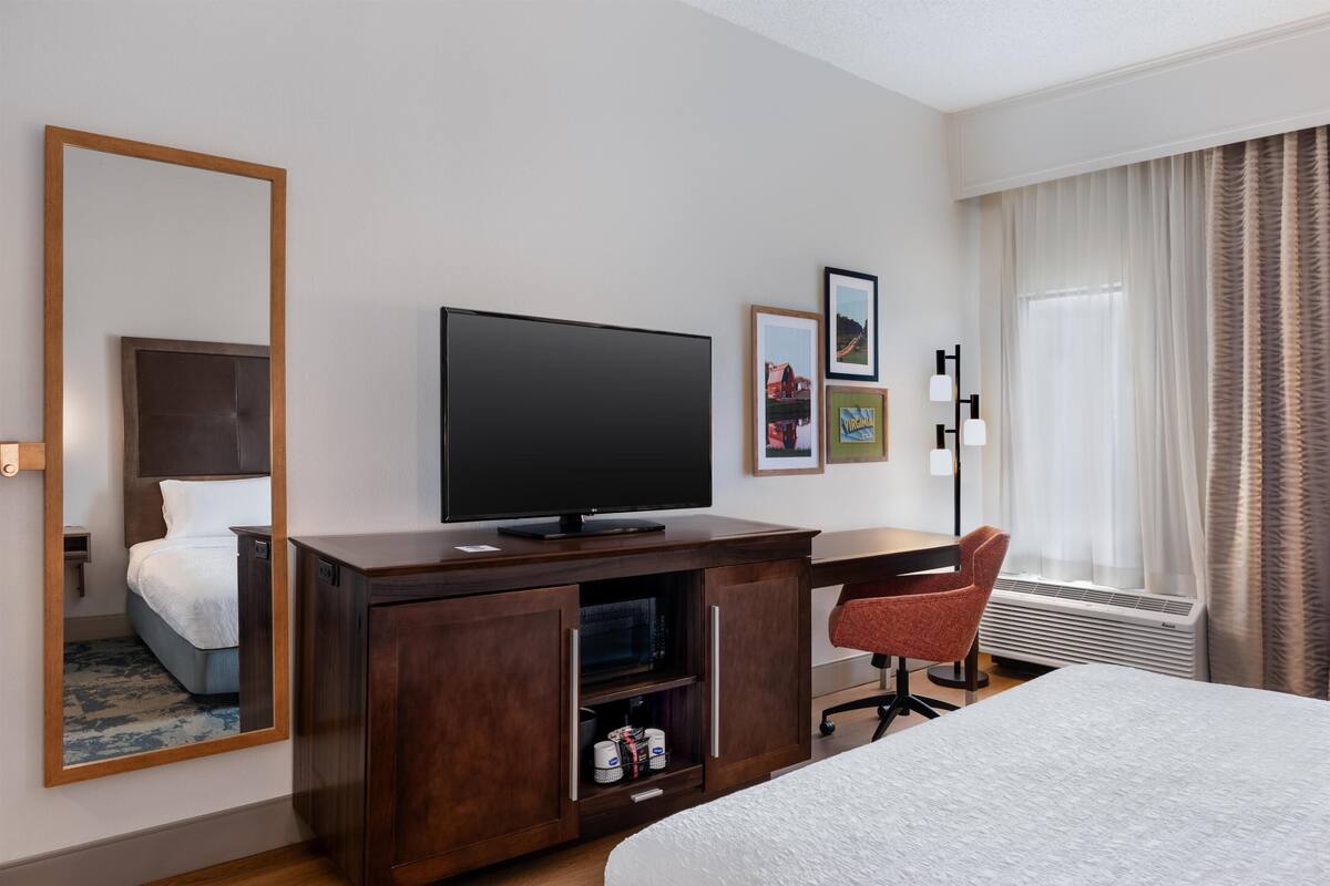 A hotel room with a bed, a TV, a desk, a chair, a mirror, and framed pictures on the wall.