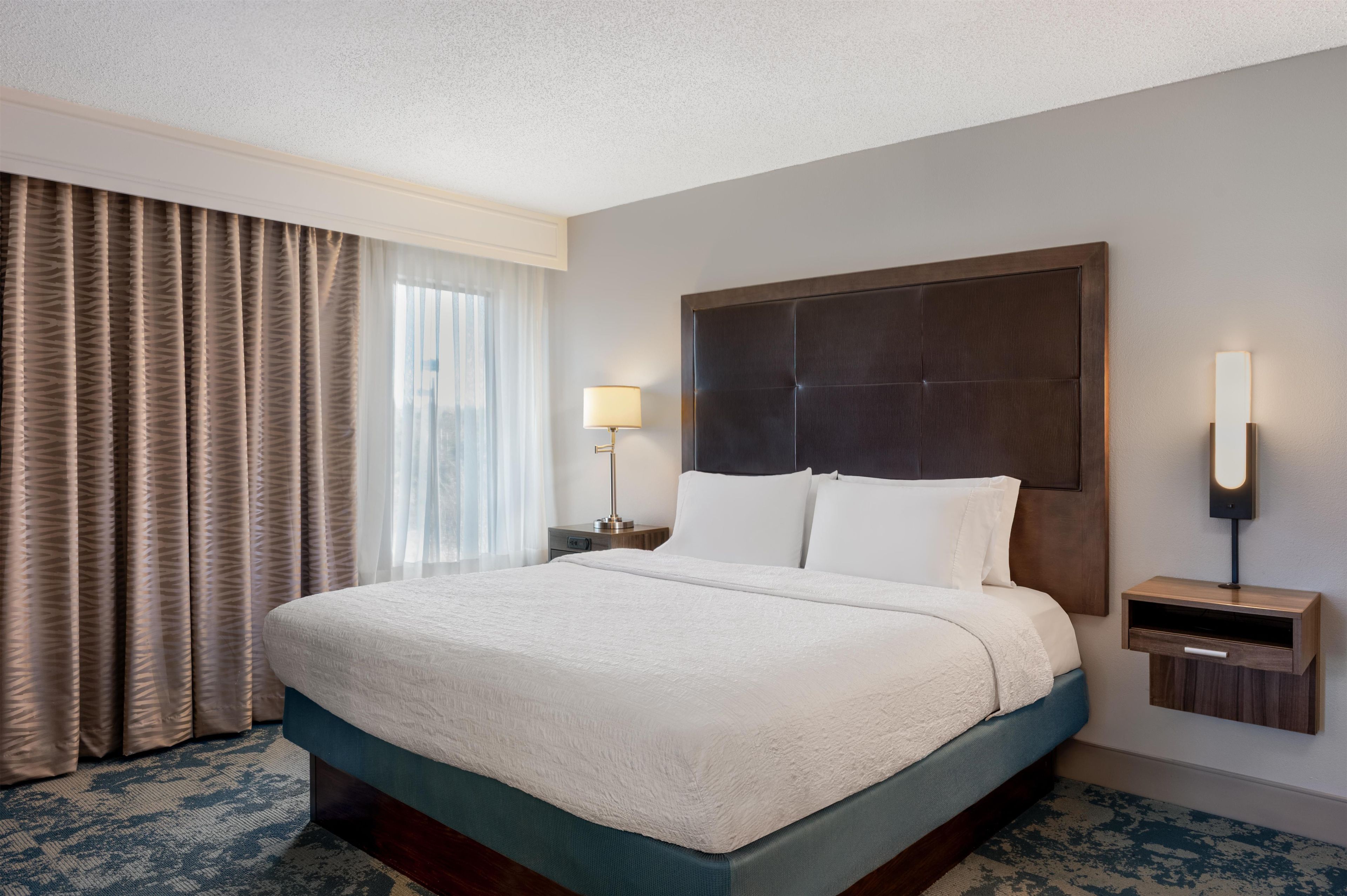 A hotel room with a large bed, a nightstand, a lamp, and a view of the city through the window.