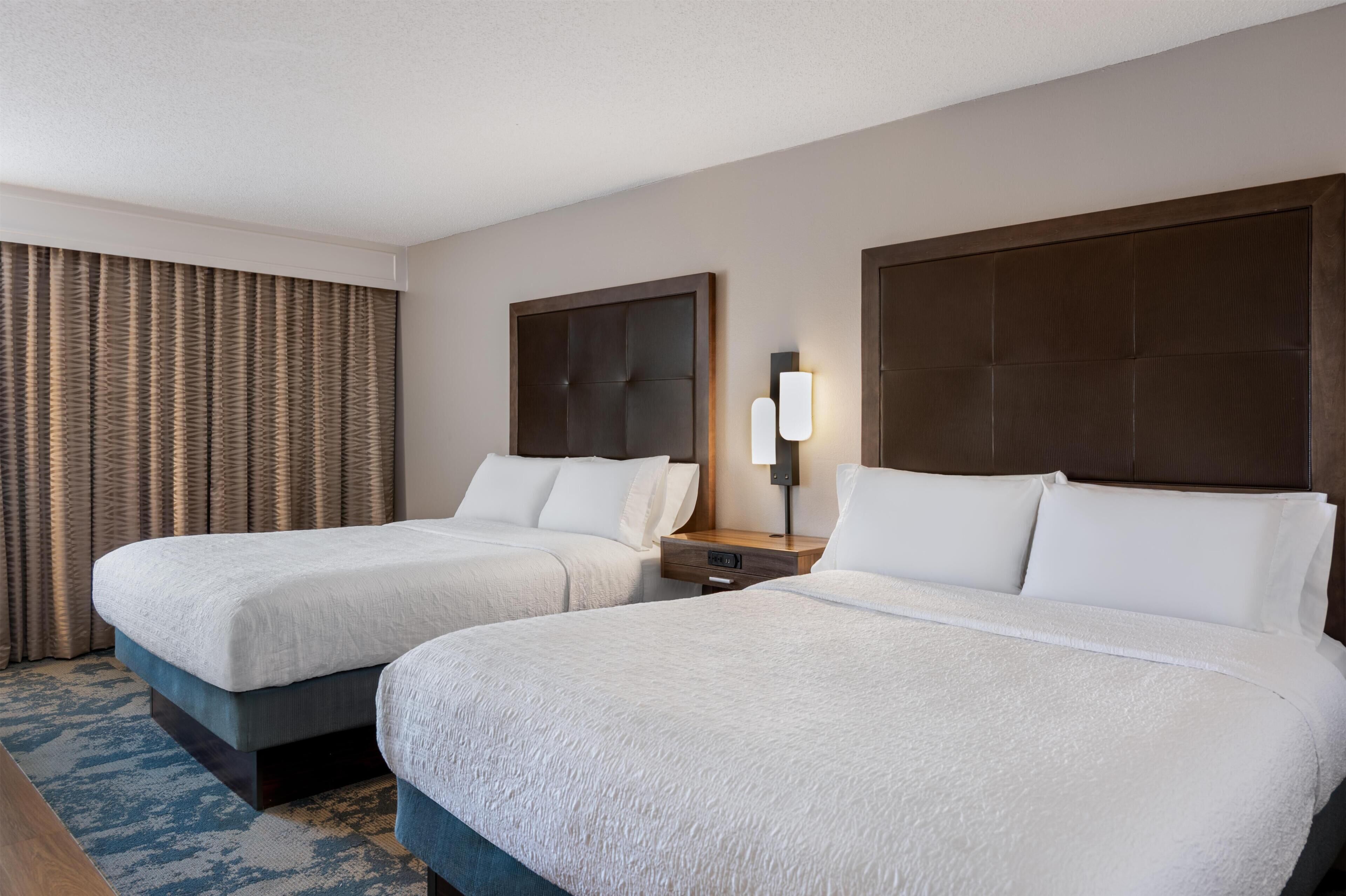 A hotel room with two beds, a nightstand, a lamp, and a large window with curtains.