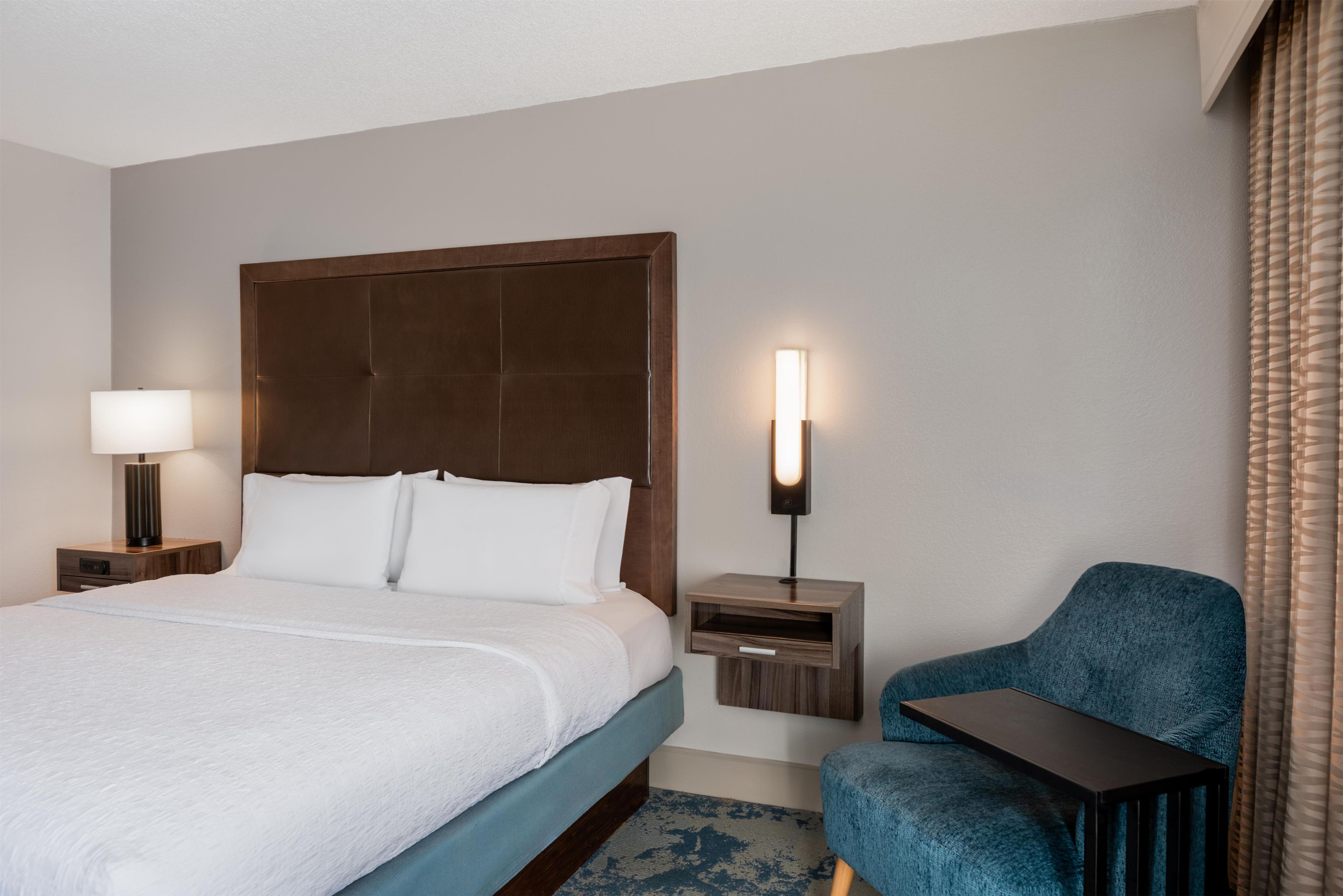 A hotel room with a large bed, a bedside table with a lamp, a chair, and a small table.