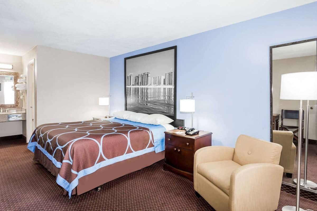 A hotel room with a bed, a nightstand, a chair, a desk, a lamp, a mirror, and a cityscape painting on the wall.
