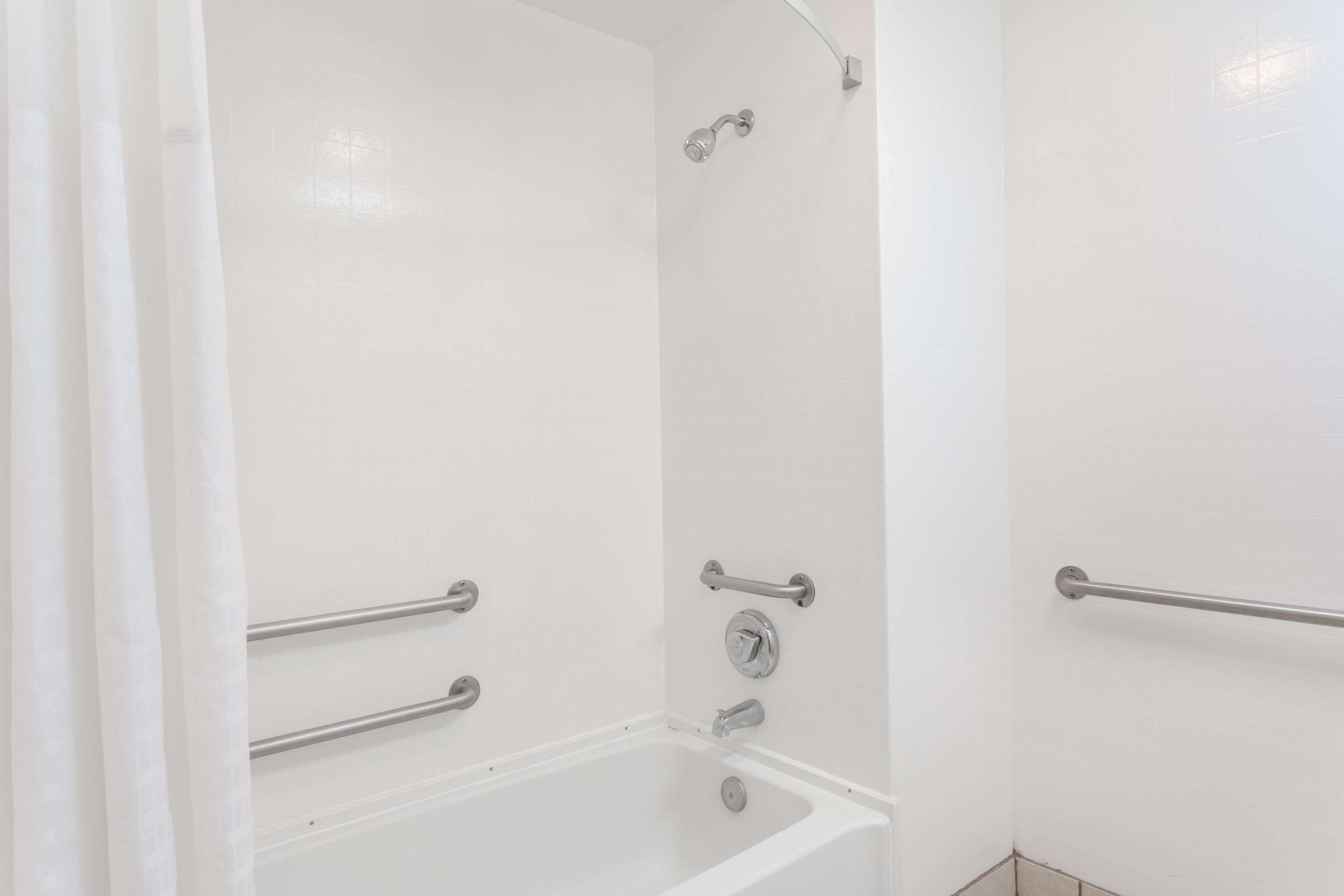Un bagno bianco con vasca, maniglie di sostegno e soffione doccia.