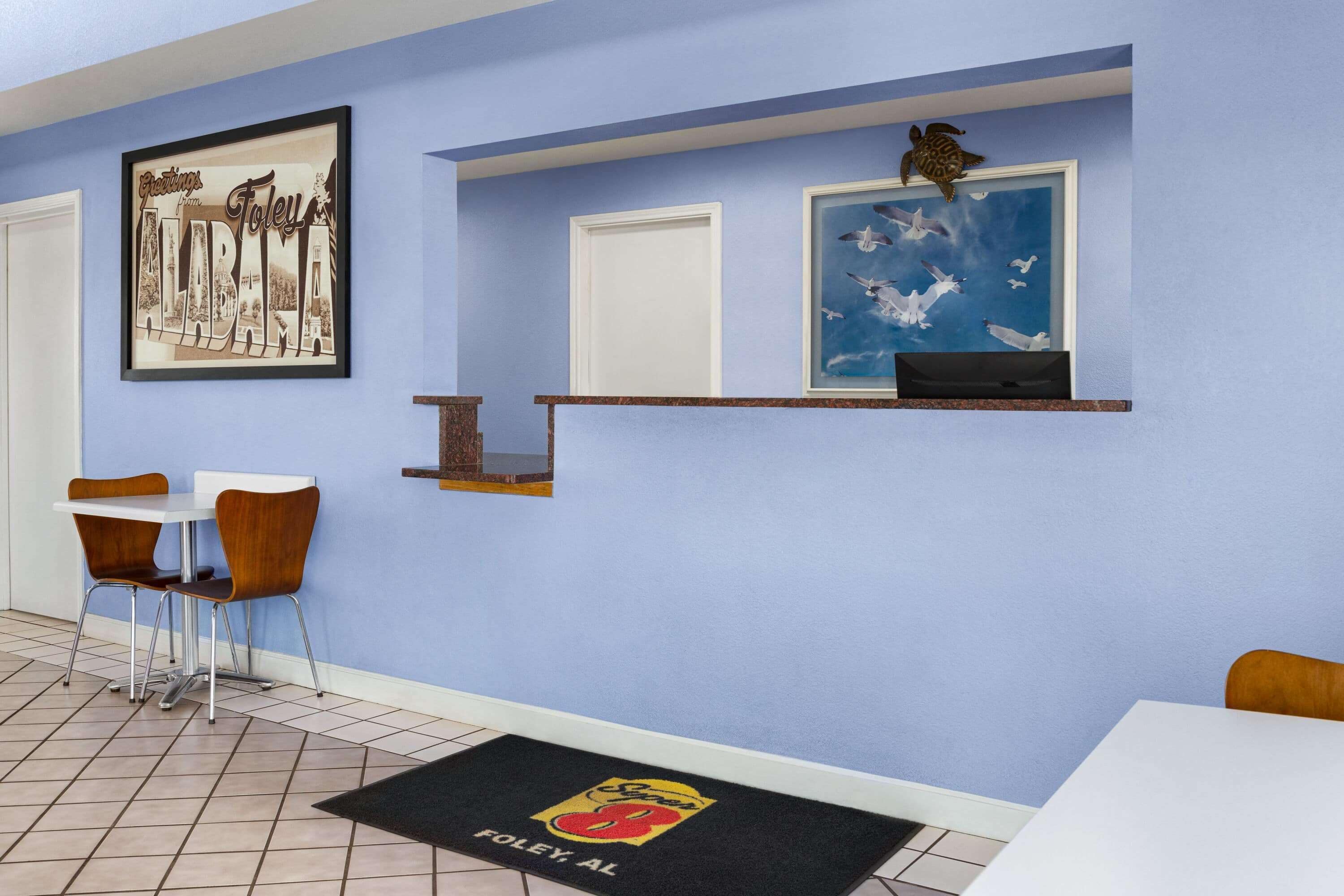 Uma área de jantar com parede azul, um letreiro de boas-vindas emoldurado, uma prateleira com uma foto emoldurada e um tapete de entrada com um logotipo.