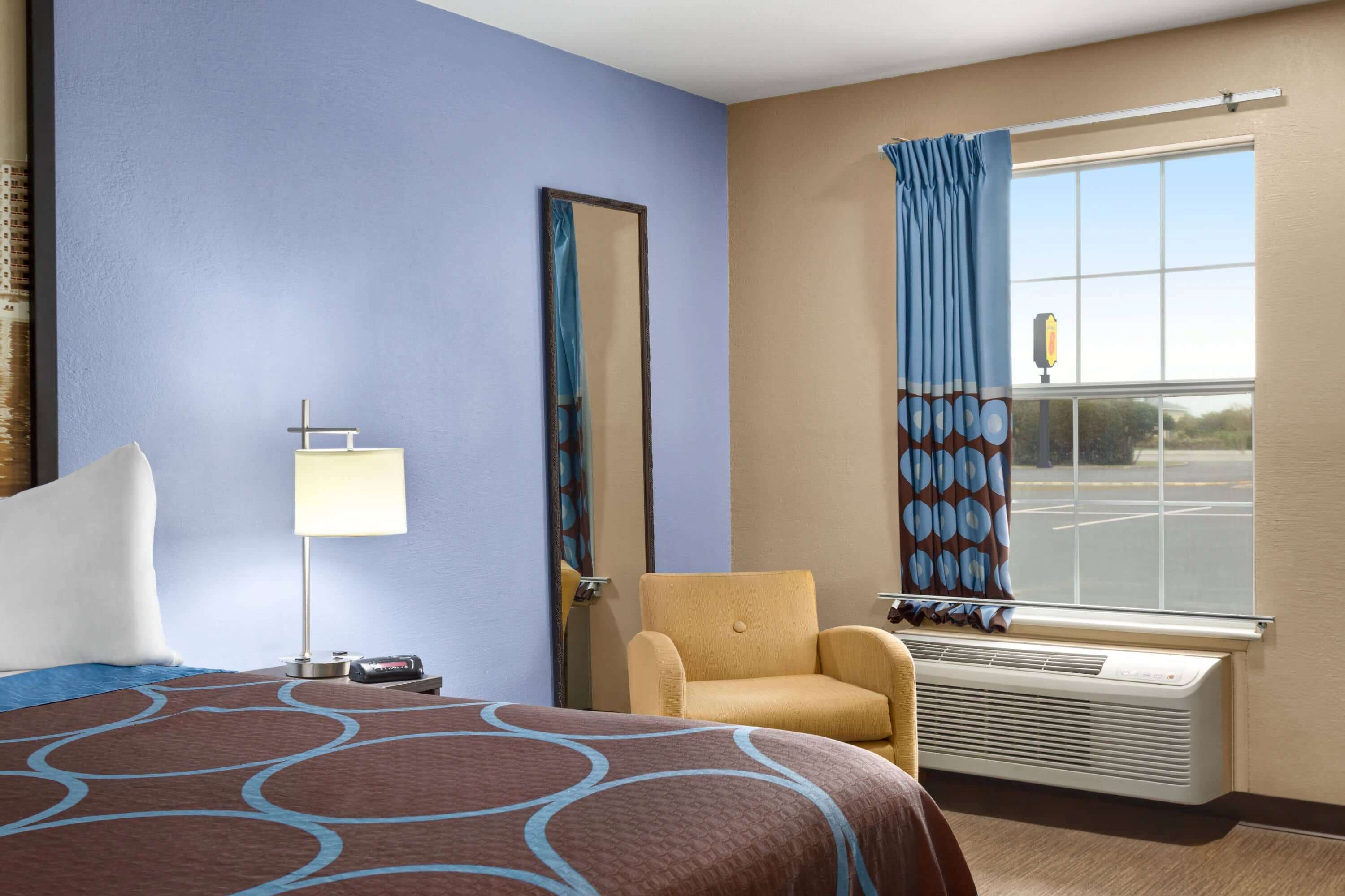Ein Hotelzimmer mit Bett, Sessel, Fenster mit Vorhängen, Lampe und Klimaanlage.