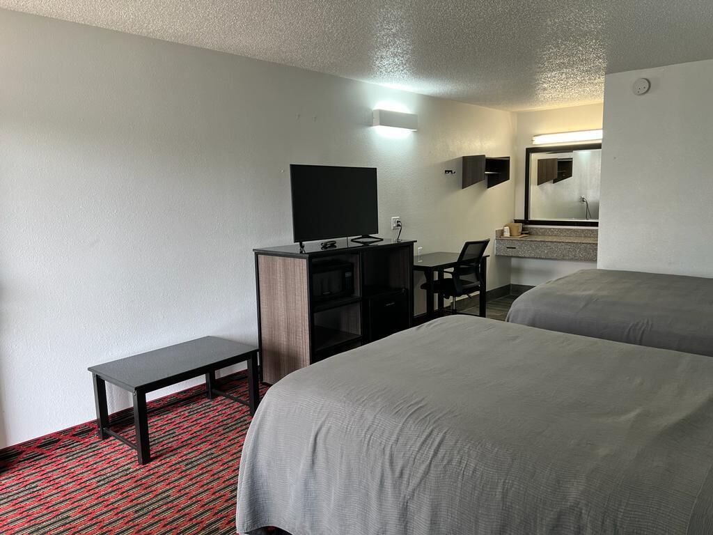 A hotel room with two beds, a desk with a flat-screen TV, a small bench, and a mirror above the desk.