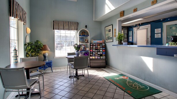 A reception area with a reception desk, a seating area with chairs and a table, a plant, a lamp, a bulletin board, and a window with a view of the outside. - Keywest Inn - Tuscumbia/Muscle Shoals (Tuscumbia)