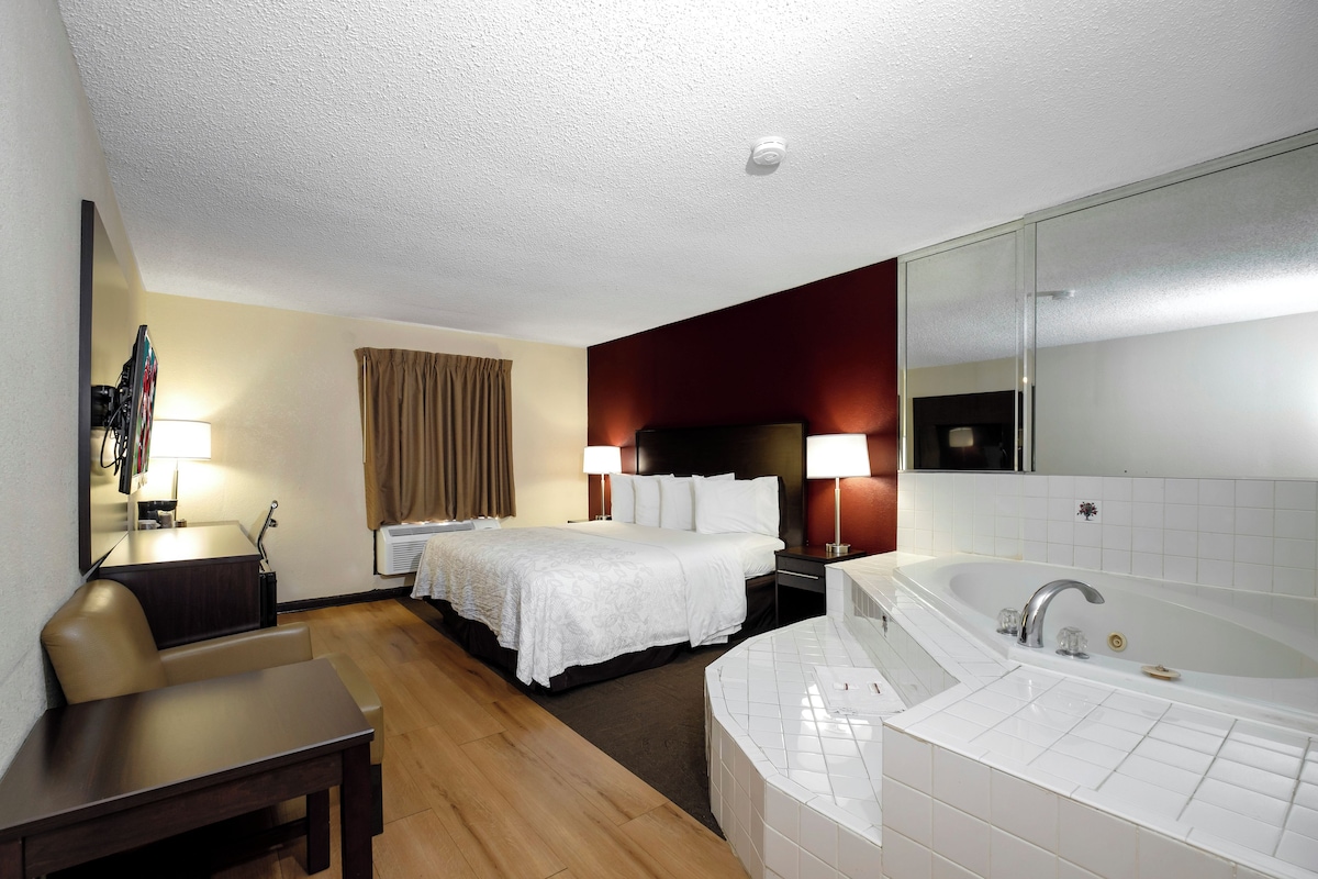 A hotel room with a bed, a desk, a chair, a large mirror, a bathroom with a bathtub, and a window with curtains.