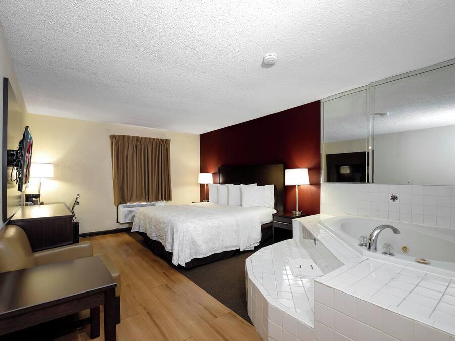 Een hotelkamer met een bed, bureau, stoel, grote spiegel, badkamer met bad en raam met gordijnen.