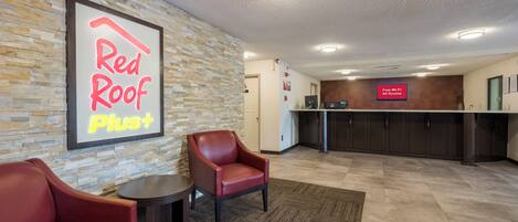 A Red Roof Plus hotel lobby with a stone wall, a reception desk, and leather chairs.