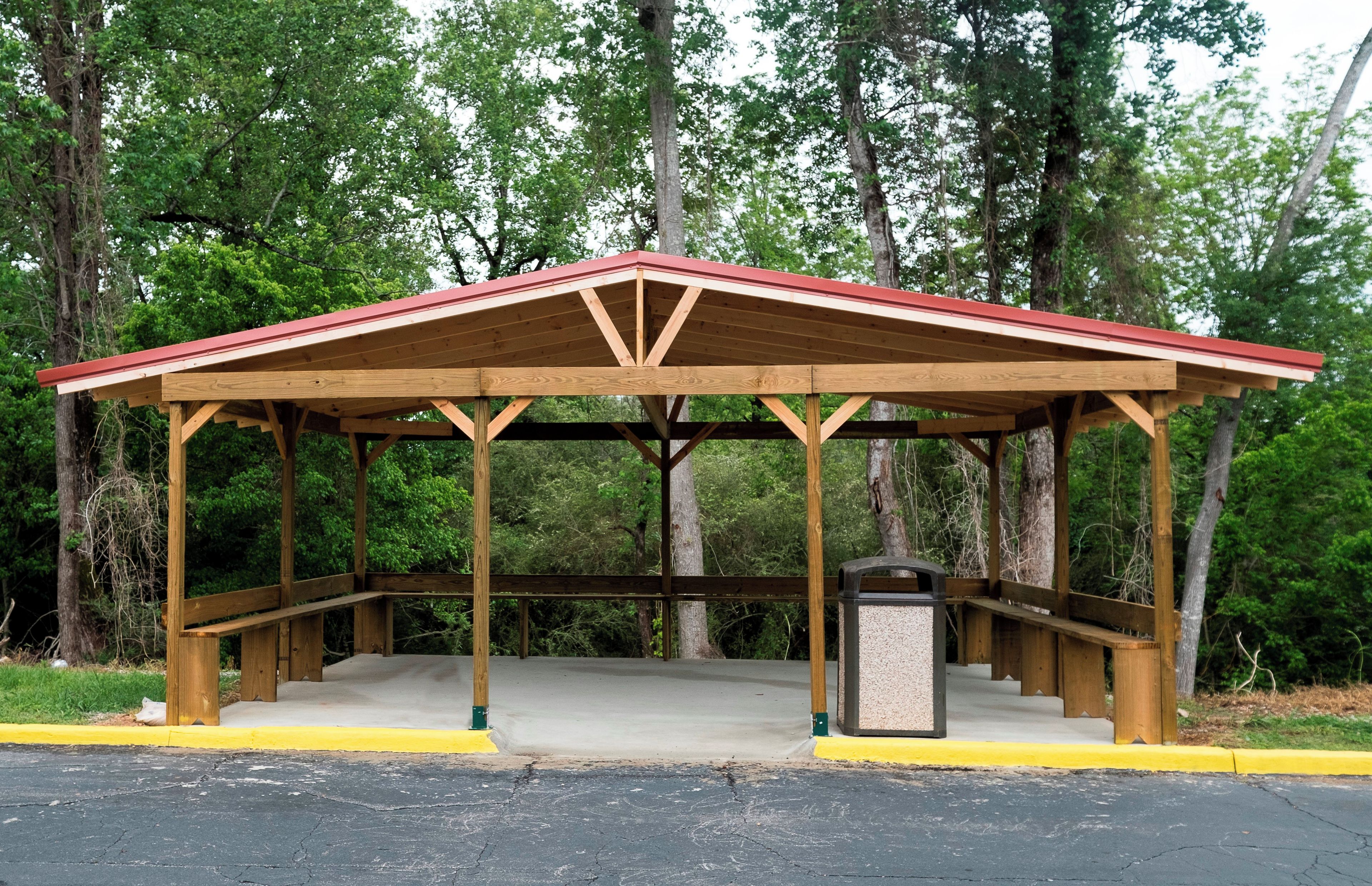 A wooden pavilion with a red roof, a concrete floor, and a trash can.