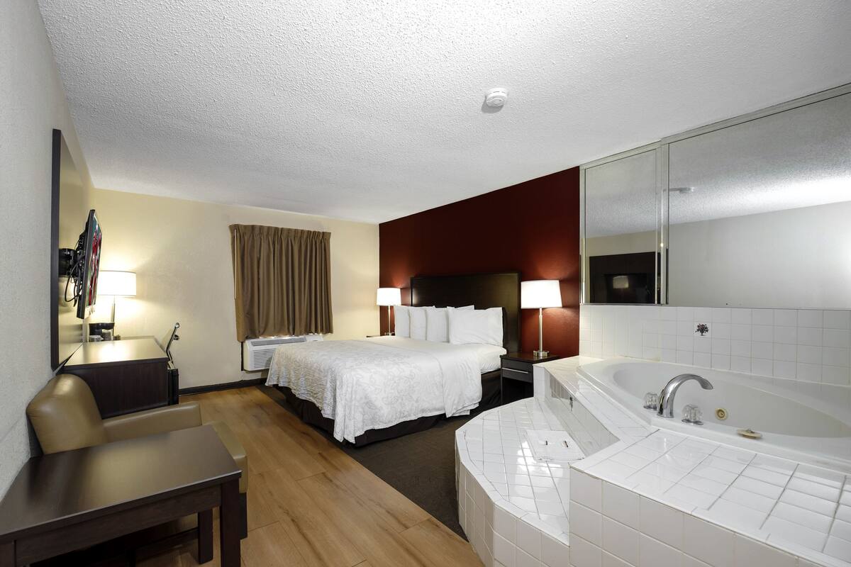 A hotel room with a bed, a desk, a chair, a large mirror, a bathtub, and a window with curtains.