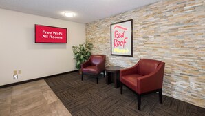 A hotel lobby with a flat-screen TV displaying 'Free Wi-Fi All Rooms,' a 'Red Roof Plus+' sign, two leather chairs, a small table, and a potted plant. - Red Roof Inn PLUS+ & Suites Opelika (Opelika)