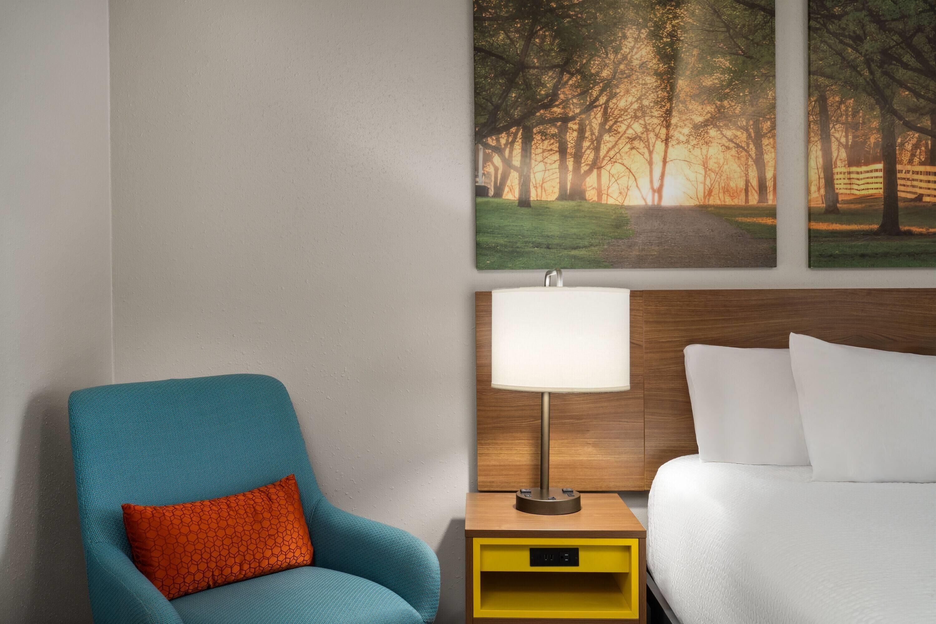 Ein Hotelzimmer mit einem Bett, einem Nachttisch mit Lampe, einem Sessel mit Kissen und zwei Wandbildern.
