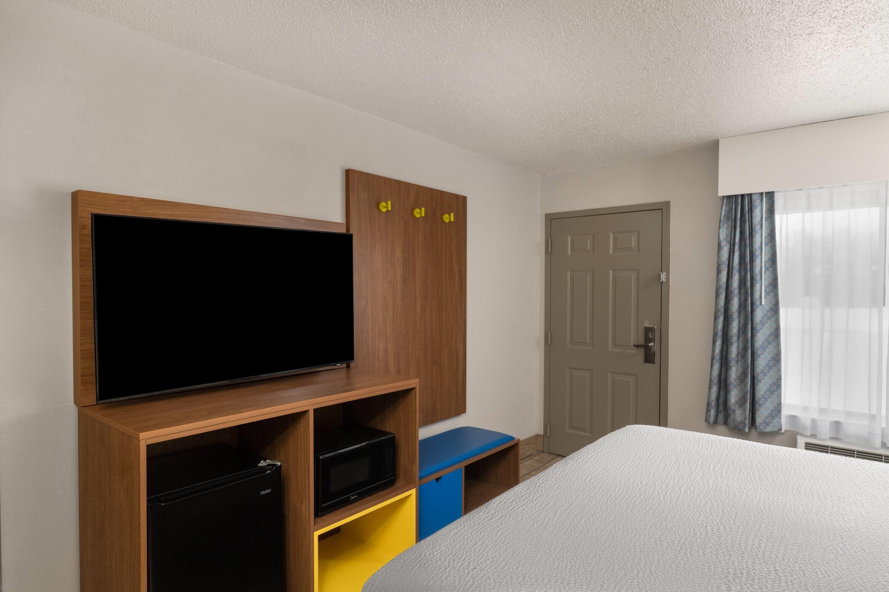 A hotel room with a bed, a television on a wooden stand, a door, and a window with curtains.
